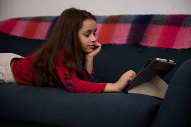 istock Adorable elementary age kid, cute little kid girl using digital tablet, watching movies while lying on the sofa at cozy home interior. Distance learning. Online education. Digital gadget addiction 2150967972