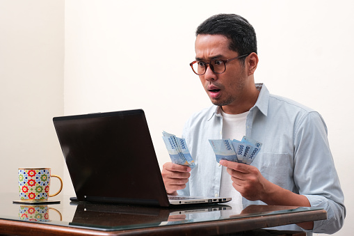 A man looking to his laptop showing shocked expression while counting money
