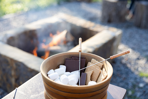 With a campfire billowing in the background, there is a bowl with ingredients to make smores.