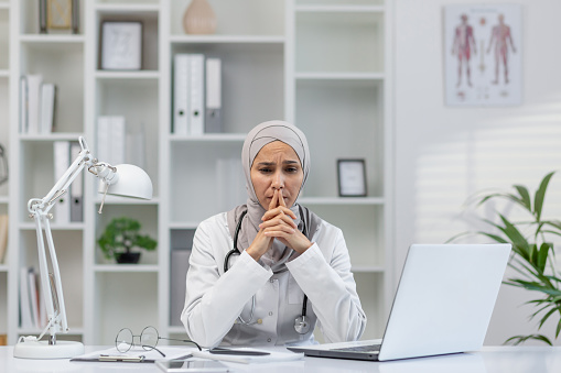 A female doctor wearing a hijab looks thoughtful and concerned while sitting at her desk in a modern clinic office, surrounded by medical books and a laptop.