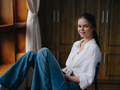 Woman sitting at home by a wooden window with a smile on a chair in homemade comfortable clothes and looking at the landscape, spring mood, women's day, rest on the weekend. High quality photo