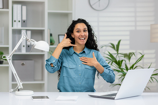 Joyful businesswoman with curly hair in a blue shirt sits at her office desk, gesturing a 'call me' sign and smiling energetically.
