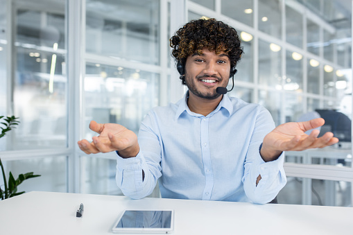 Young male customer service representative in office setting, wearing headset, smiling and gesturing warmly. Modern, bright workplace environment captured.
