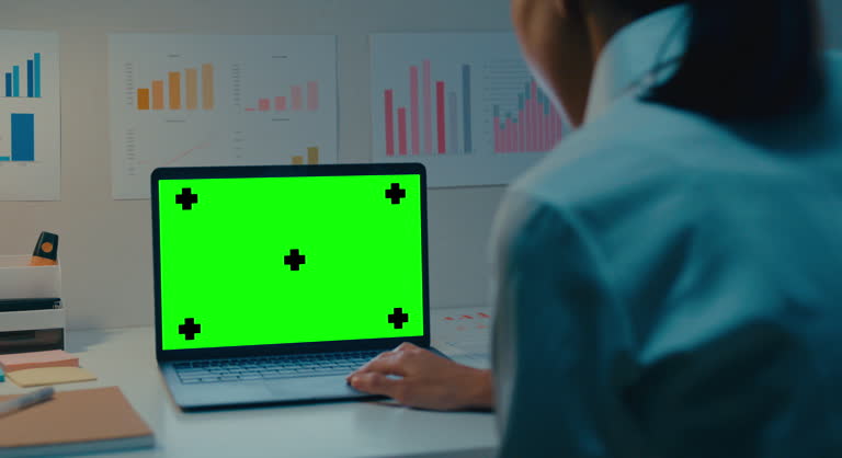 Closeup of young Asian businesswoman using laptop with mock-up green screen while working in modern office at night. Chroma key technology, Marketing design.
