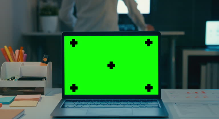 Closeup of laptop with mock-up green screen standing on desk in the modern creative office at night. Chroma key technology, Marketing design.