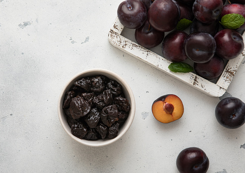 Sweet prunes in glass bowl with ripe plums in wooden box on light kitchen background.