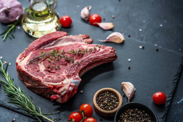 istock Raw beef steak on the bone on a black slate serving plate, decorated with various ingredients such as cooking oil, dry pepper, cherry tomatoes and herbs. 2150997177