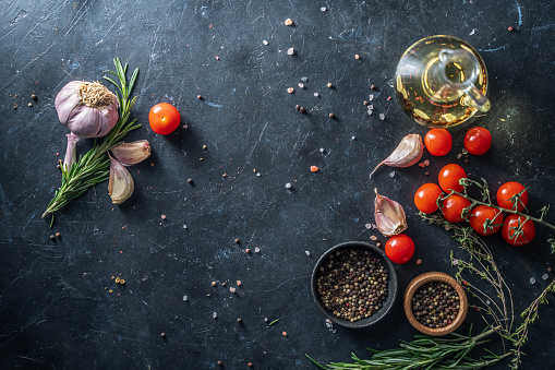 Decanter of cooking oil, tomatoes, rosemary, garlic and mixed dry pepper on black rough background.