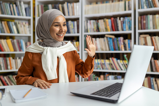 A cheerful Muslim woman wearing a hijab uses a laptop in a library setting, waving and smiling, exuding friendliness and accessibility while surrounded by books.