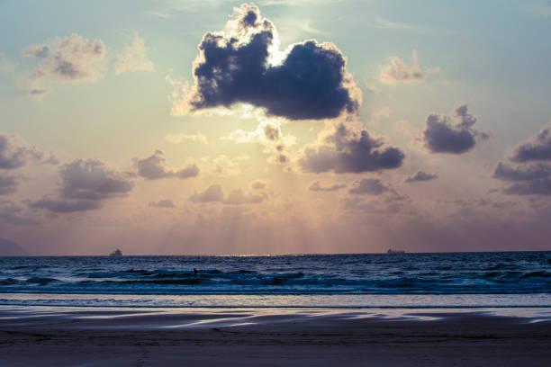 A large cloud is in the sky above the ocean. The sun is setting, casting a warm glow over the water. The sky is filled with clouds, creating a moody atmosphere