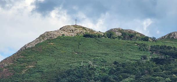 This awe-inspiring photograph captures the majestic peak of a lush green hill, crowned with a solitary cross that stands as a poignant symbol against the cloudy sky. The hillside, richly carpeted with varying shades of green and dotted with patches of purple heather, suggests a serene yet rugged natural landscape. The cross at the summit not only enhances the spiritual allure of the scene but also invites viewers to contemplate the peaceful solitude that such heights can offer.