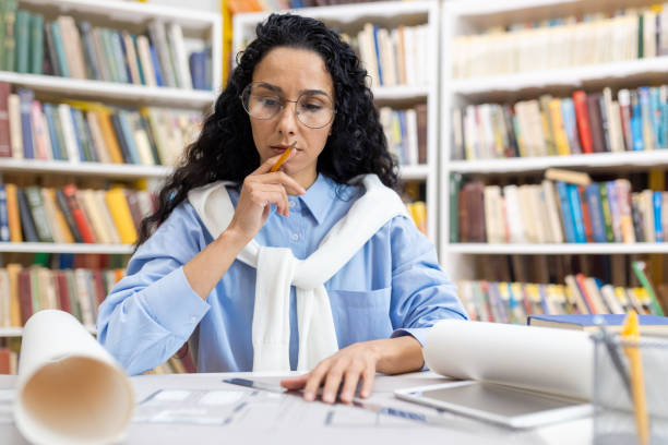 istock Focused female architect reviewing blueprints in library 2151015040