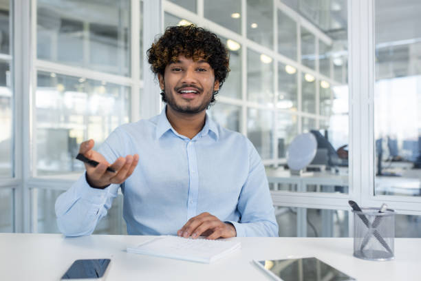 istock Indian male spreading hands with pen to camera while sitting in front of table with notebook. Smiling hr manager asking question from candidates for hiring and making remarks in copybook 2151016505