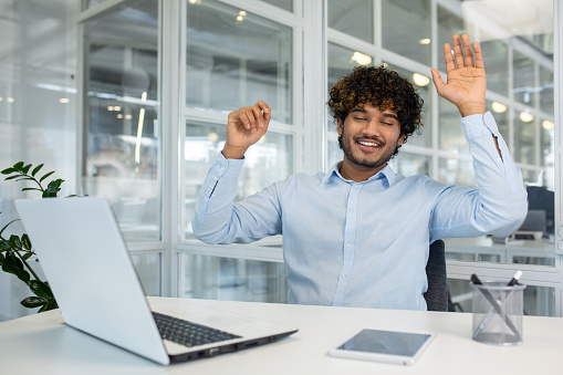 Curly islamic man in blue shirt raising arms for stretching back while sitting at workspace with closed eyes. Relaxed employee using free time for exercising and resting from sedentary work.