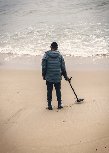 Cascais, Portugal - April 20, 2024: A lone unidentifiable man stands on a sandy beach, holding a metal detector as he scans the shoreline for hidden treasures. He wears a winter jacket and jeans
