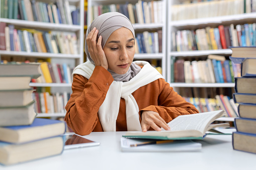 A thoughtful Muslim woman in a hijab immersed in her studies at a library, surrounded by piles of books, showcasing dedication and the pursuit of knowledge.