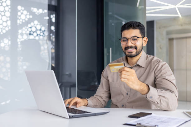 istock Positive unshaved male in eyewear looking at debit card while keeping hand on keyboard of modern computer. Carefree office worker enjoying possibility of making online payments in marketplaces 2151020779
