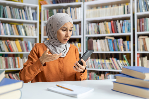 A young Muslim woman wearing a hijab expresses frustration while trying to solve a problem using her smartphone in a library setting surrounded by books.