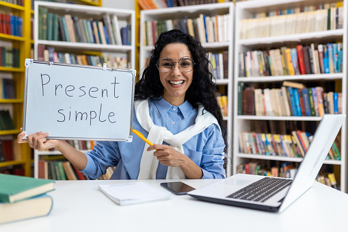 Cheerful female teacher holding a board with 'Present Simple' written on it, demonstrating English grammar in a library setting surrounded by books.