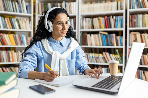 A female student wearing headphones concentrates on her laptop while studying in a library. She is surrounded by books and has coffee nearby, indicating a long study session.