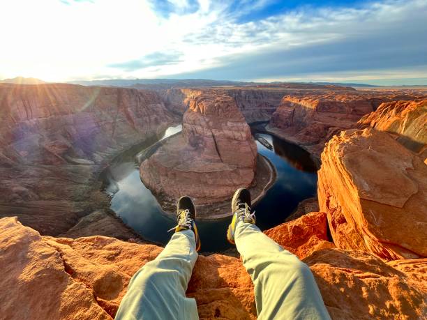 POV of young adult man sitting in front of Horseshoe Bend, Arizona, United States of America. POV Point of view of young adult man sitting in front of Horseshoe Bend while enjoying his own free time and rest looking the landscape view at top of Grand Canyon National Park in Arizona, United States Of America at sunset in winter time.
He's wearing green pants and black boots. horseshoe bend river stock pictures, royalty-free photos & images