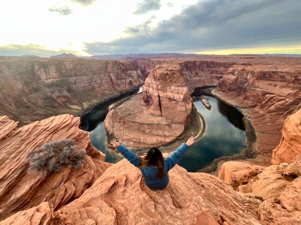 An adult traveler backpacker female open her arms in sign of freedom while sitting on top of Horseshoe Bend while rest and enjoying the landscape view at Glen / Grand Canyon National Park in Arizona, United States Of America. An adult traveler backpacker female open her arms in sign of freedom while sitting on top of Horseshoe Bend while rest and enjoying the landscape view at Glen / Grand Canyon National Park in Arizona, United States Of America at sunset time in winter season.
She's wearing a blue winter jacket. horseshoe bend river stock pictures, royalty-free photos & images