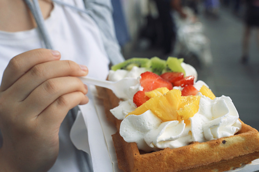 A close-up view of a hand holding a delicious waffle topped with fresh strawberries, kiwi, and a dollop of whipped cream, ready to be enjoyed at an outdoor setting.