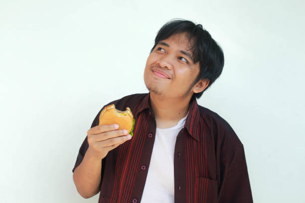 A young Asian man with a thinking and happy expression holds and eats a beef burger. Isolated in gray background.