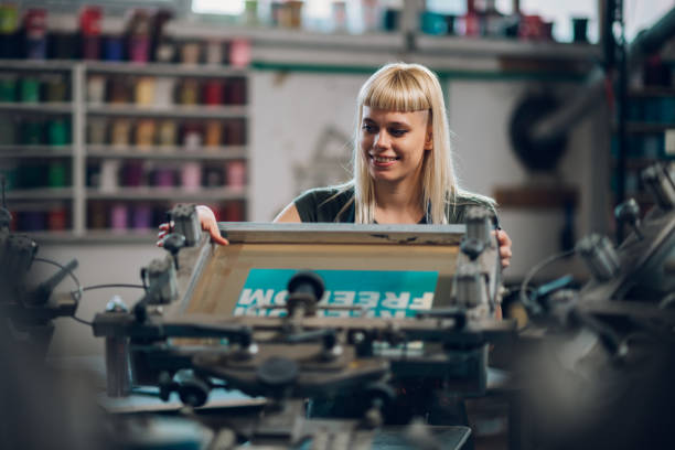 istock Female graphic worker putting screen printing plate on printing press. 2151033678