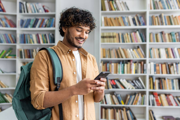 istock Positive middle eastern man wearing backpack slung over one shoulder standing in front of bookshelf and holding modern mobile phone. Happy male chatting with friends online in social medias 2151036110