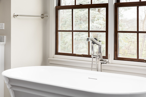 A freestanding bathtub faucet detail in front of a wood framed window with a blurred ourside and tan walls.