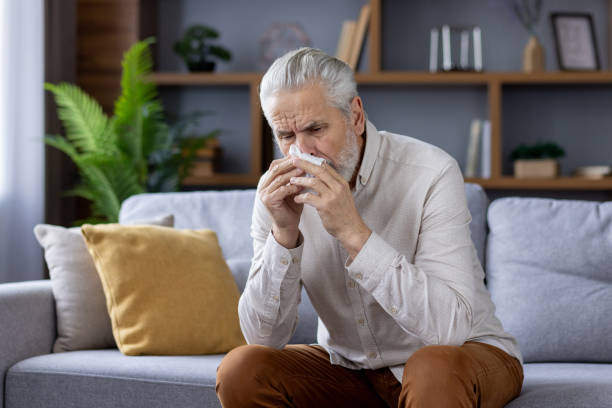 istock Senior man with a cold sitting and blowing nose at home 2151040313
