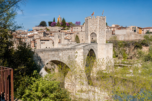 The picturesque medieval bridge in the village of Besalú, Girona, crosses the river to access the village in the background. The bridge is old and has a rustic look.