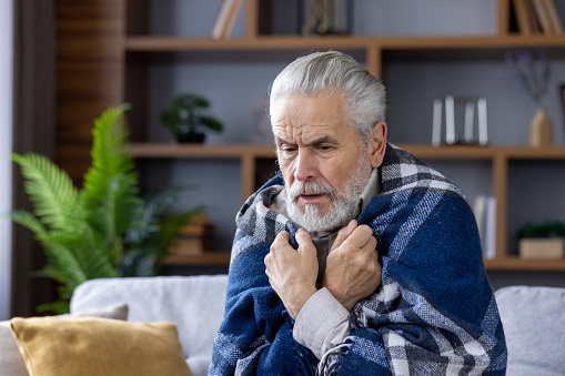 A close-up photo of a gray-haired older man sick from the virus, who is sitting at home on the sofa covered with a blanket and shivering from the cold and temperature.