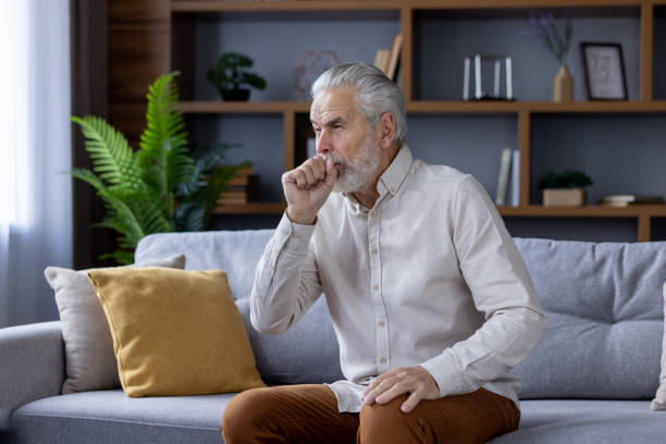 istock Senior man sitting on sofa coughing in modern living room 2151044357