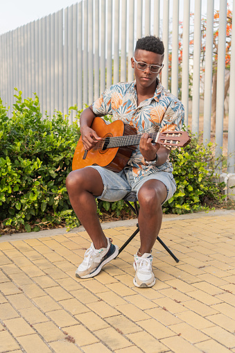 A young African man in sunglasses and a vibrant tropical shirt plays an acoustic guitar while sitting on a stool outdoors.
