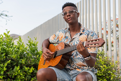 Young musician with eyeglasses playing an acoustic guitar, surrounded by lush greenery in a modern urban environment.