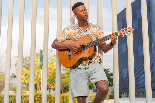 Focused African man playing guitar in park, styled in a floral shirt and denim shorts, with urban architecture and greenery background.