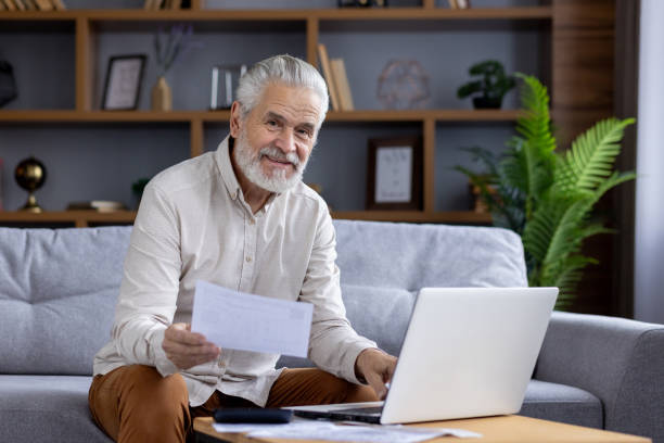 istock Senior man smiling while working with laptop and documents at home 2151044689