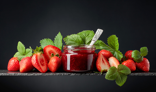Strawberry jam in a small jar and fresh berries with leaves on a black background.
