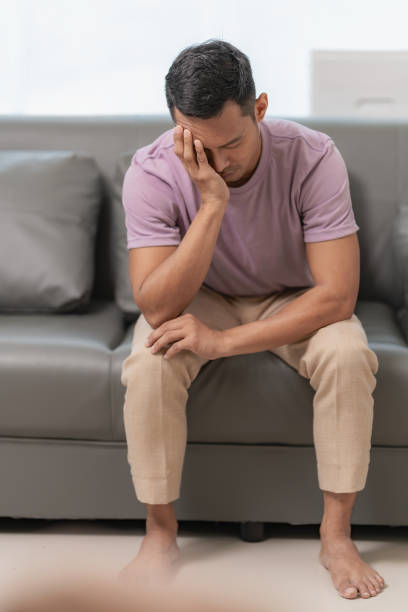 istock Sad man suffering from a headache or migraine touching his temples who is massaging his head, stressed sitting on the sofa with a pained expression on his face. Feeling weak or depressed 2151061246