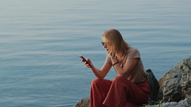 Relaxed blonde woman using her mobile phone on the tranquil sea background. Internet surfing outdoors.