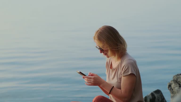 Relaxed blonde woman using her mobile phone on the tranquil sea background. Internet surfing outdoors.