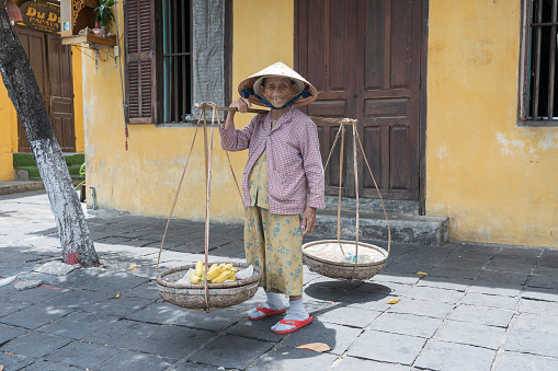 Hoi An, Vietnam - July 01, 2020 : Vietnamese woman in a straw hat with a basket of fruits on a street market in the old city against the background of the old yellow wall in Hoi An, Vietnam
