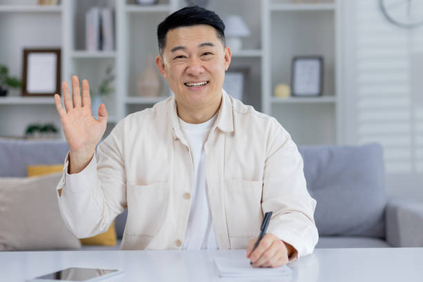 istock Smiling Asian man waving while seated at home 2151072947
