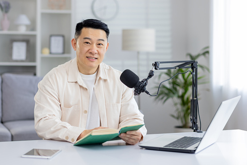 A focused man in a stylish, bright home office setup engrossed in a book with a podcasting microphone mounted on the desk.