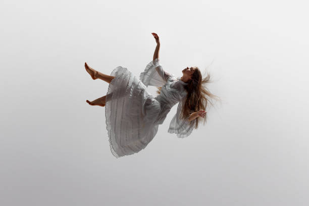 Woman in white ruffled dress floats effortlessly in moment of graceful falling in mid-air against white studio background. Woman in white ruffled dress floats effortlessly in moment of graceful falling in mid-air against white studio background. Concept of beauty, feminine elegance and purity, dream and reality. floating woman stock pictures, royalty-free photos & images