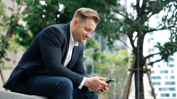 istock Businessman sitting at park while working on phone by reading report. Urbane 2151083883