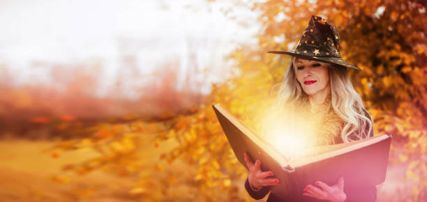 Halloween a woman with a magic spell book in a witch hat is conjuring, performing witchcraft over the autumn background of the forest.