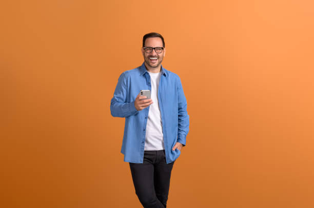 istock Cheerful young man with hand in pocket using smart phone and posing confidently on orange background 2151088073